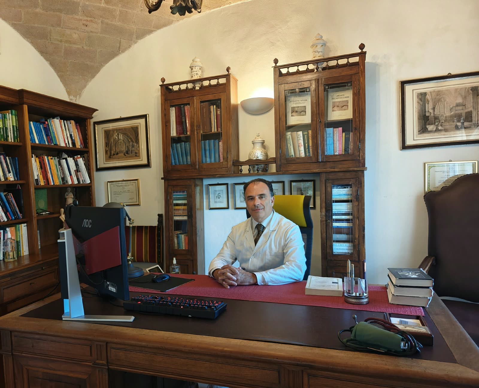 Il Dott. Tancredi Ascani in camice bianco alla scrivania del suo studio, davanti a vetrine in legno con libri e antichi vasi da farmacia.