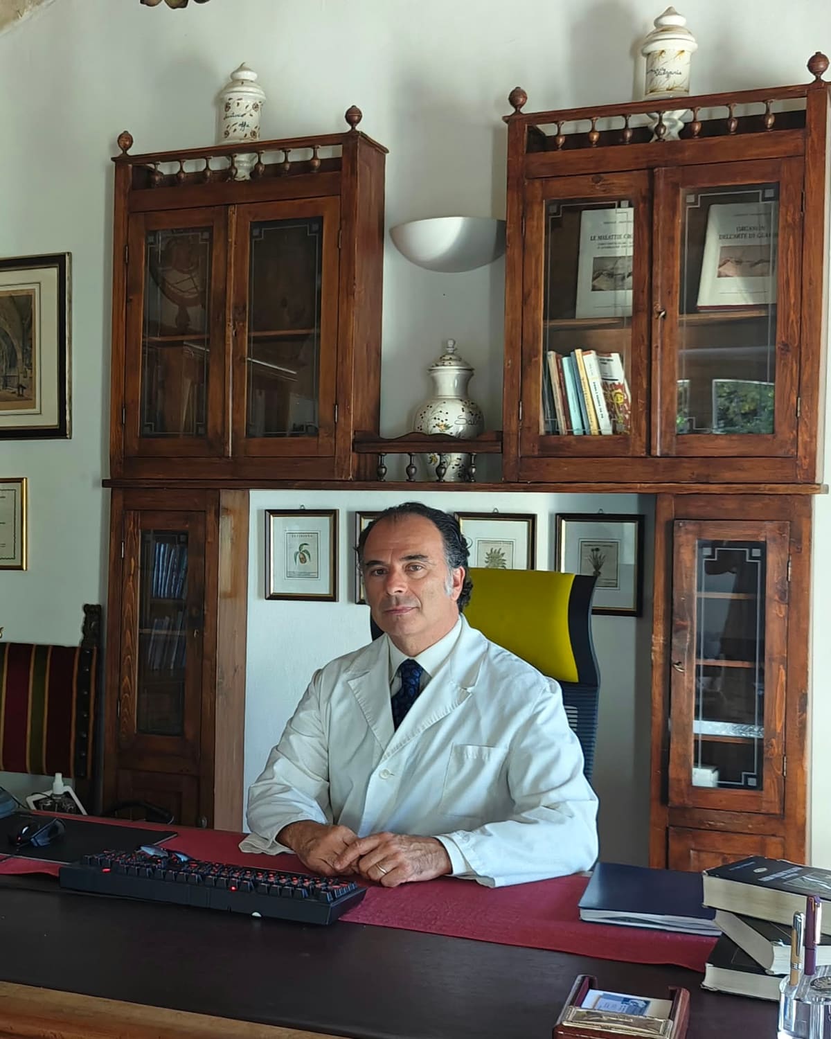 Il Dott. Tancredi Ascani in camice bianco alla scrivania del suo studio, davanti a vetrine in legno con libri e antichi vasi da farmacia.